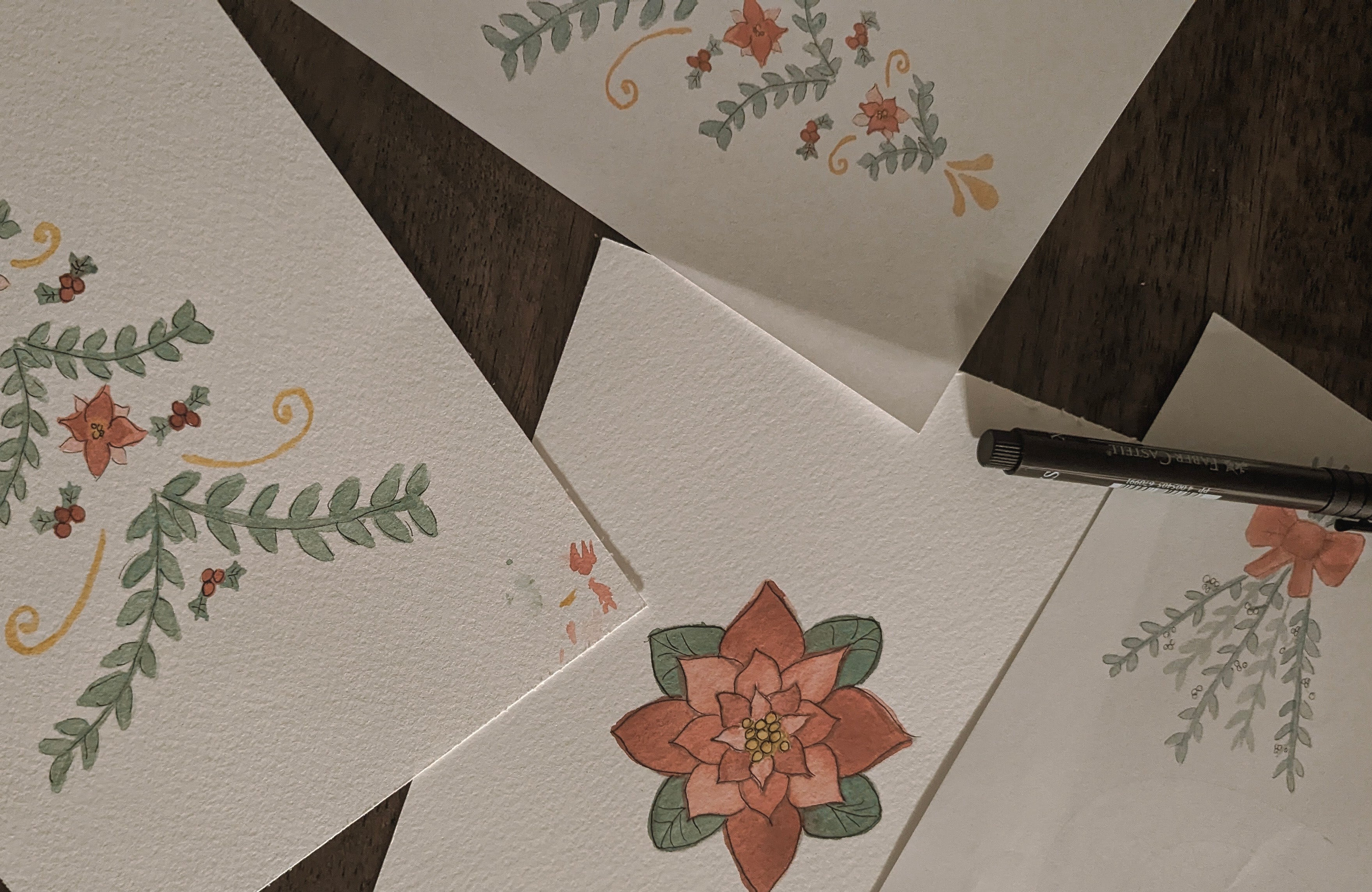 watercolor paintings of a poinsettia flower, folk inspired christmas tree, and mistletoe tied with a bow lay spread on a table