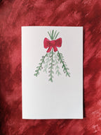 a white greeting card with a watercolor and pen drawing of branches of mistletoe tied with a red bow