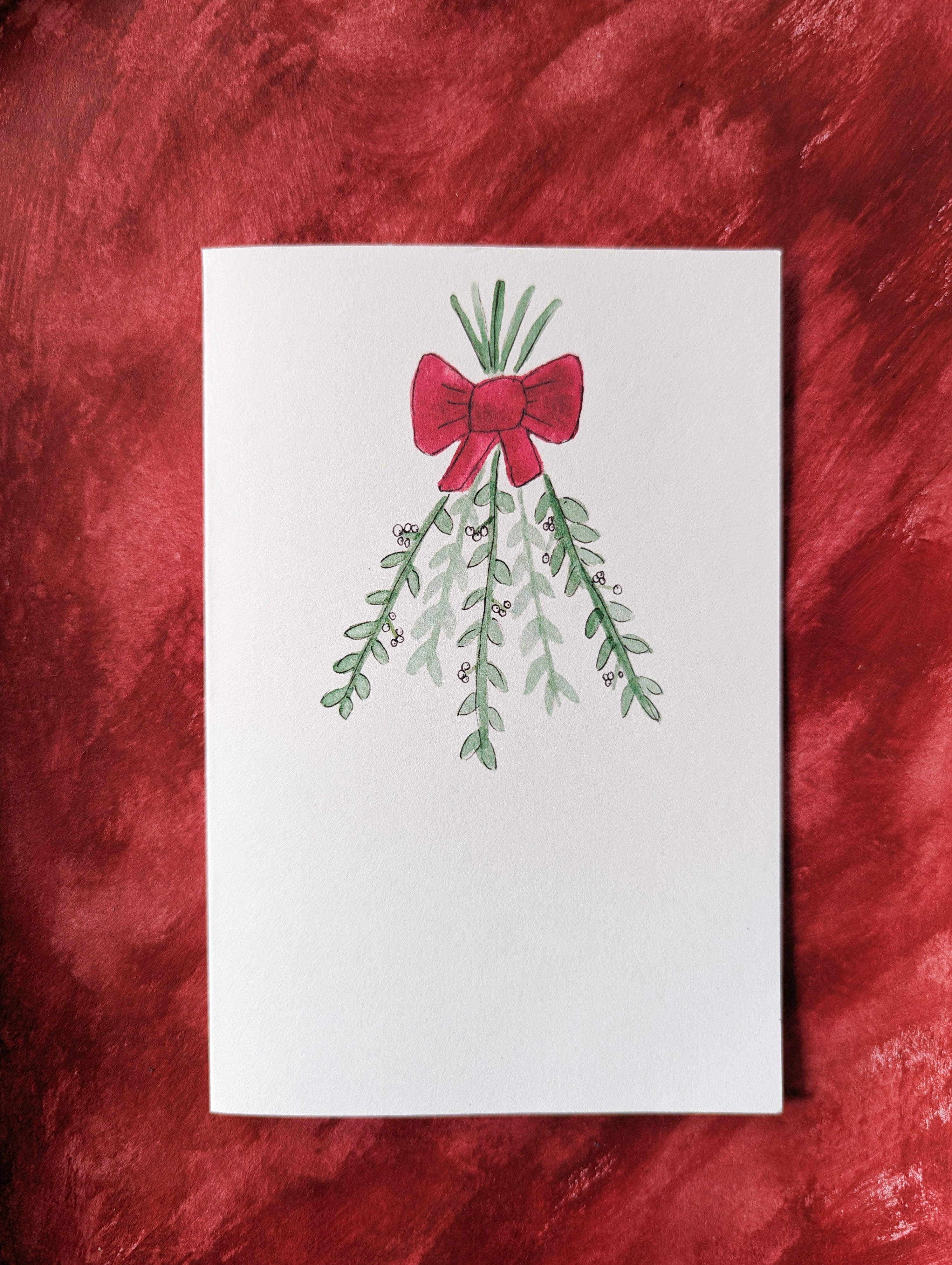 a white greeting card with a watercolor and pen drawing of branches of mistletoe tied with a red bow