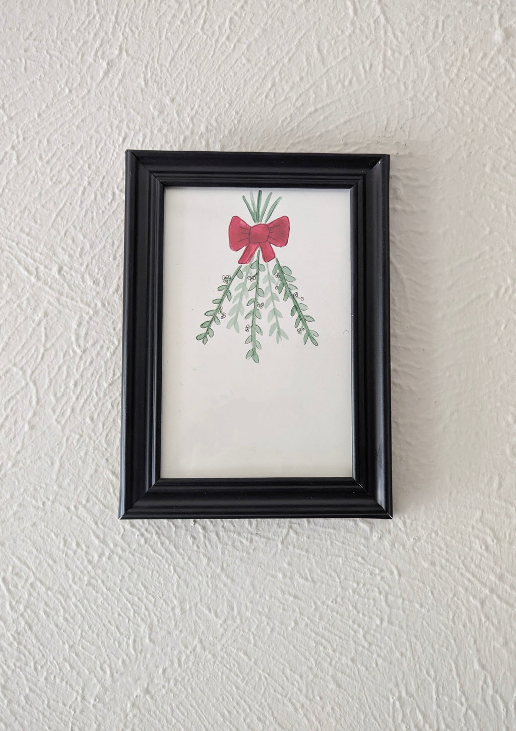a framed watercolor and pen drawing of mistletoe branches tied with a red bow hangs on a plain white wall