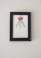 a framed watercolor and pen drawing of mistletoe branches tied with a red bow hangs on a plain white wall