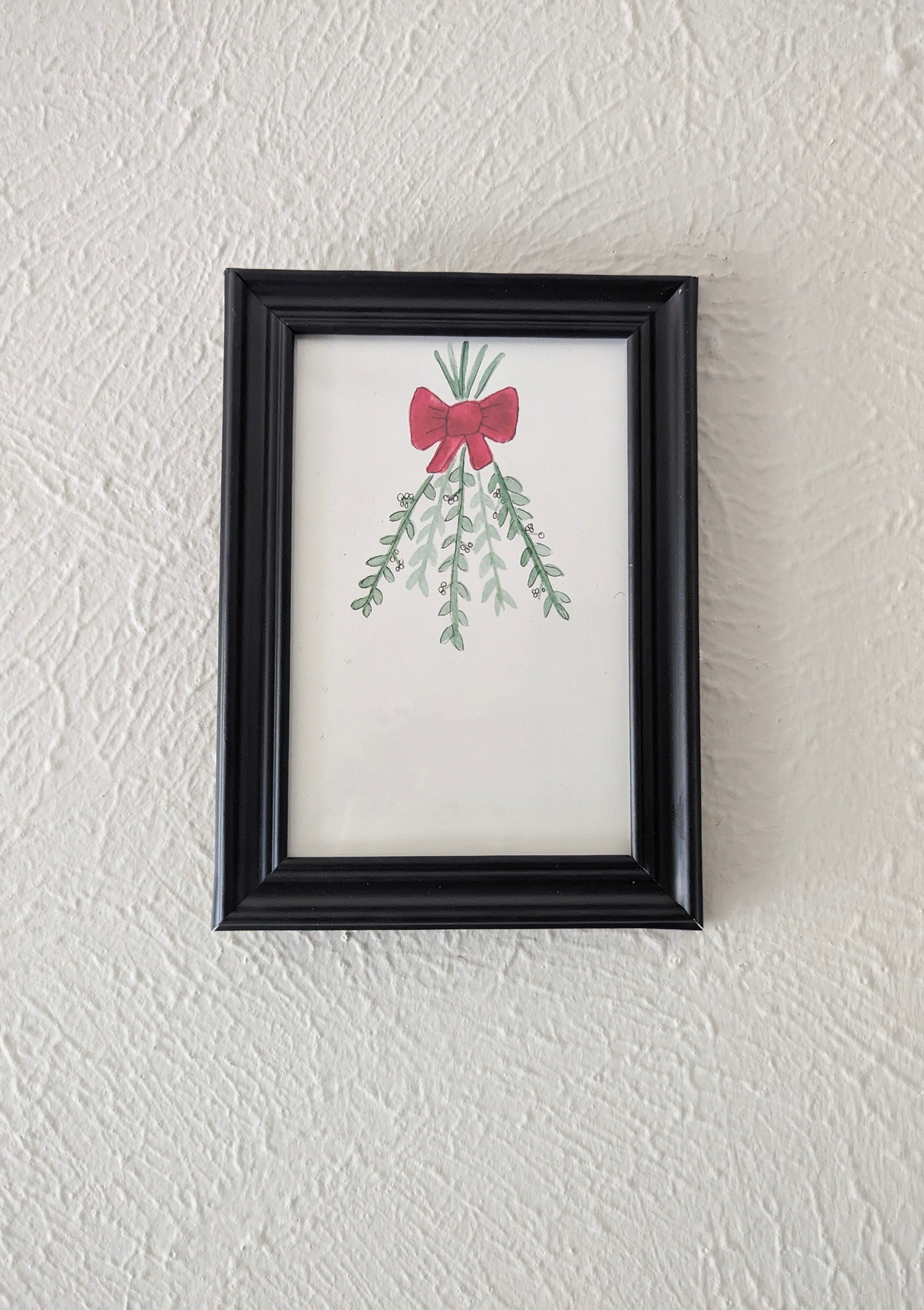 a framed watercolor and pen drawing of mistletoe branches tied with a red bow hangs on a plain white wall