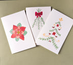 three white greeting cards with watercolor and pen drawings. One of a modern style Poinsettia flower, one of mistletoe branches tied with a red bow, and one of a folk art inspired christmas tree with greenery branches, poinsettia flowers, and holly berries